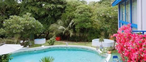 A swimming pool surrounded by lush greenery, with a building and outdoor seating area.