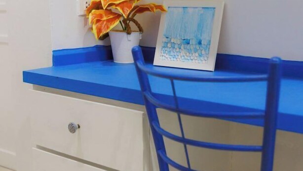 Une chaise bleue est placée devant un bureau blanc doté de tiroirs, et à côté se trouve un pot de fleurs contenant une plante aux feuilles jaunes.