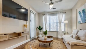 A modern living room with a flat-screen TV, a ceiling fan, a round wooden coffee table, a sofa, and a large window with blinds. - Luxury 1B w Pool near Legacy West (Frisco)