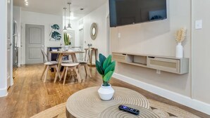 A modern living room with a dining area, wooden flooring, a mounted TV, and a wall-mounted shelf with decorative items. - Luxury 1B w Pool near Legacy West (Frisco)