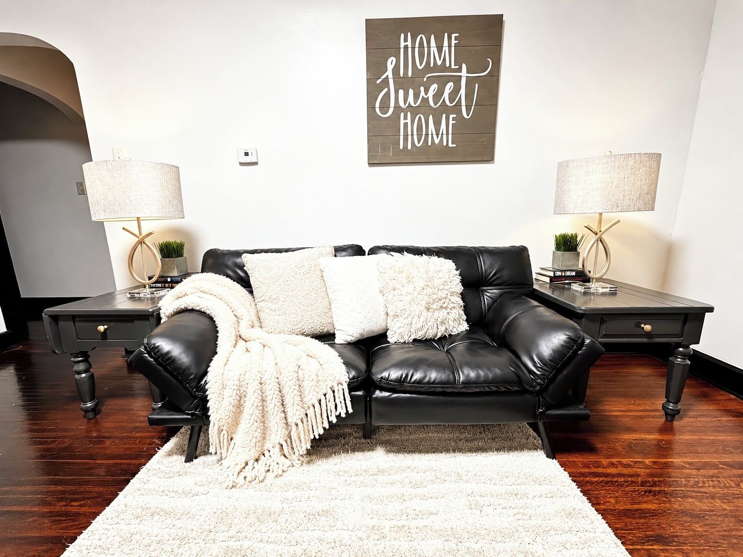 A black leather sofa with white throw pillows and a fluffy white throw blanket, flanked by two bedside tables with lamps, against a white wall with a 'HOME sweet HOME' sign.