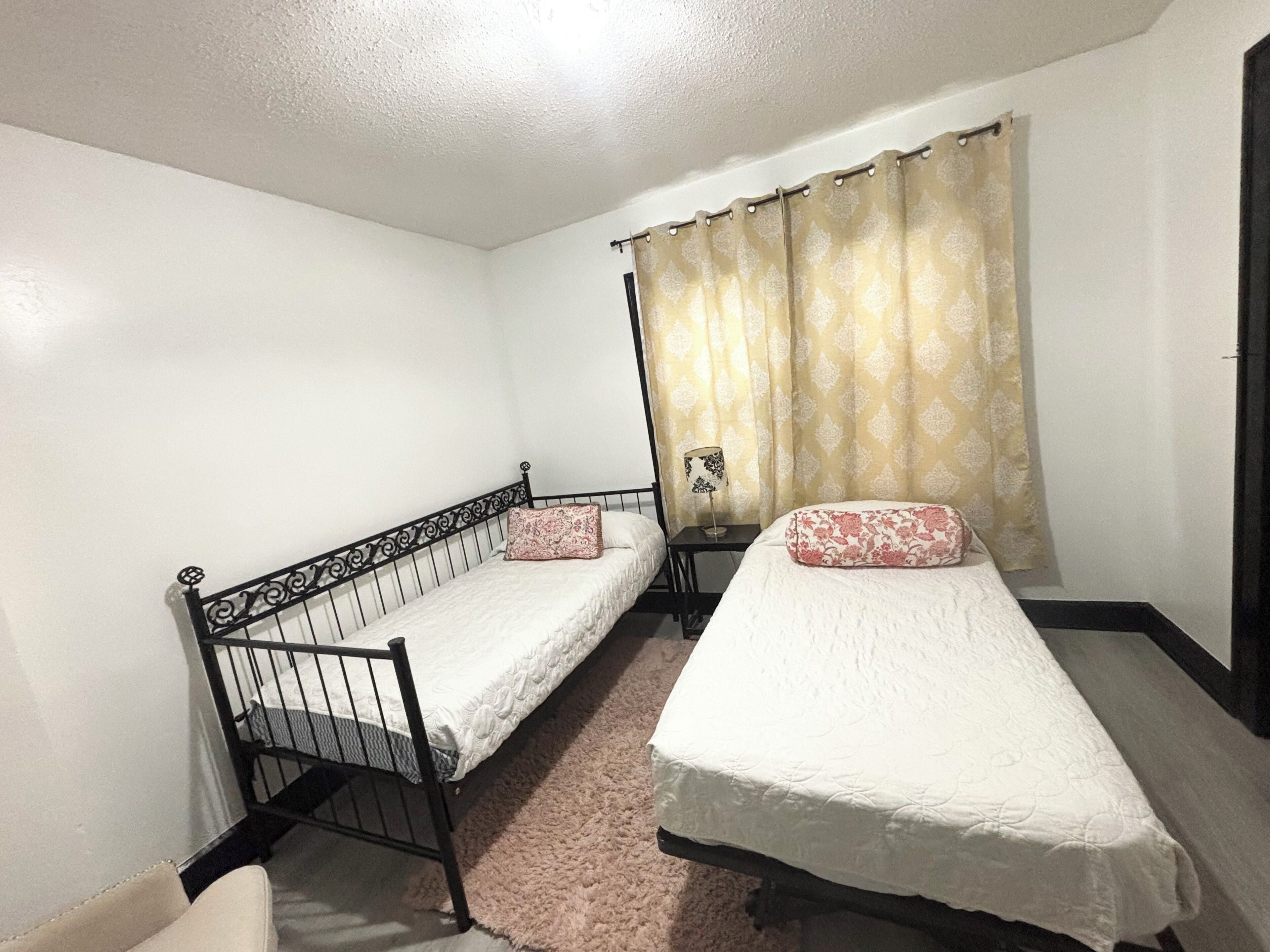 Quarto com duas camas de solteiro, uma mesa pequena e cortina.