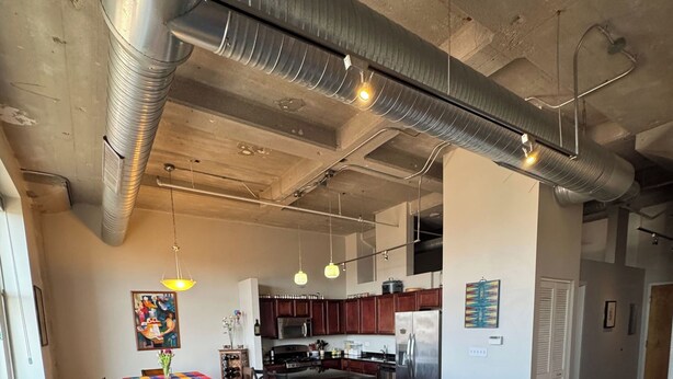 Una sala moderna con un sofá modular, una mesa de centro y una alfombra. Se observa una cocina al fondo con electrodomésticos de acero inoxidable.