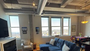 Una sala de estar moderna con un sofá modular, una mesita redonda y un televisor. Amplias ventanas ofrecen vistas a la ciudad.