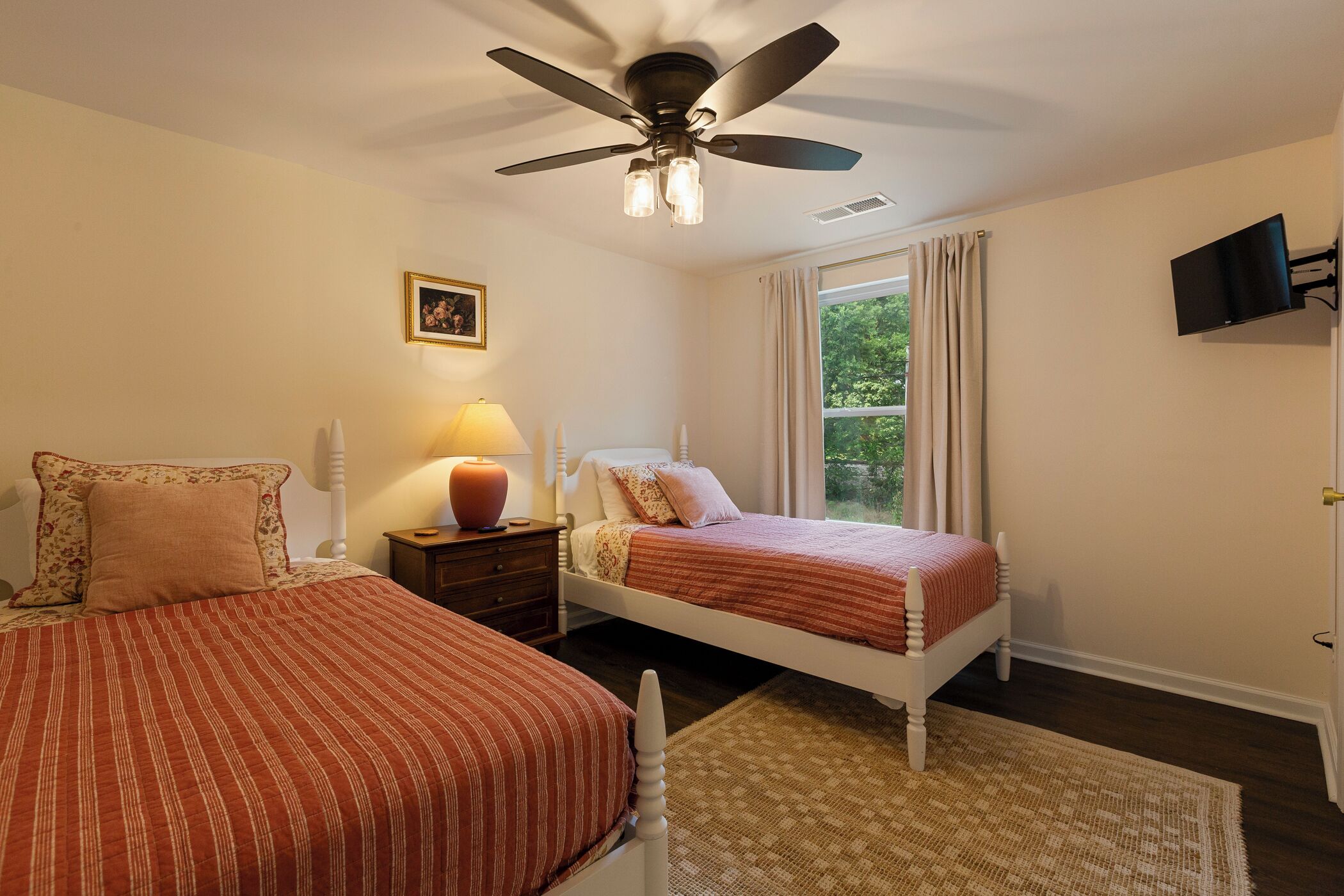 A bedroom with two beds, a ceiling fan, a bedside table with a lamp, a window with curtains, and a wall-mounted TV.