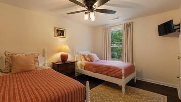 A bedroom with two beds, a ceiling fan, a bedside table with a lamp, a window with curtains, and a wall-mounted TV.