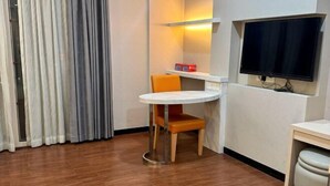 A small room with a round table, a single orange chair, a TV mounted on the wall, and a window with curtains.