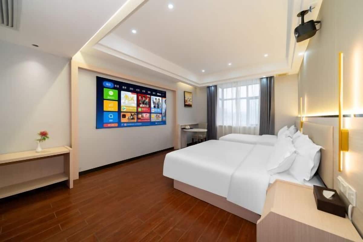 A hotel room with a large digital screen displaying various apps, a bed with white bedding, a bedside table with a lamp, and a window with curtains.