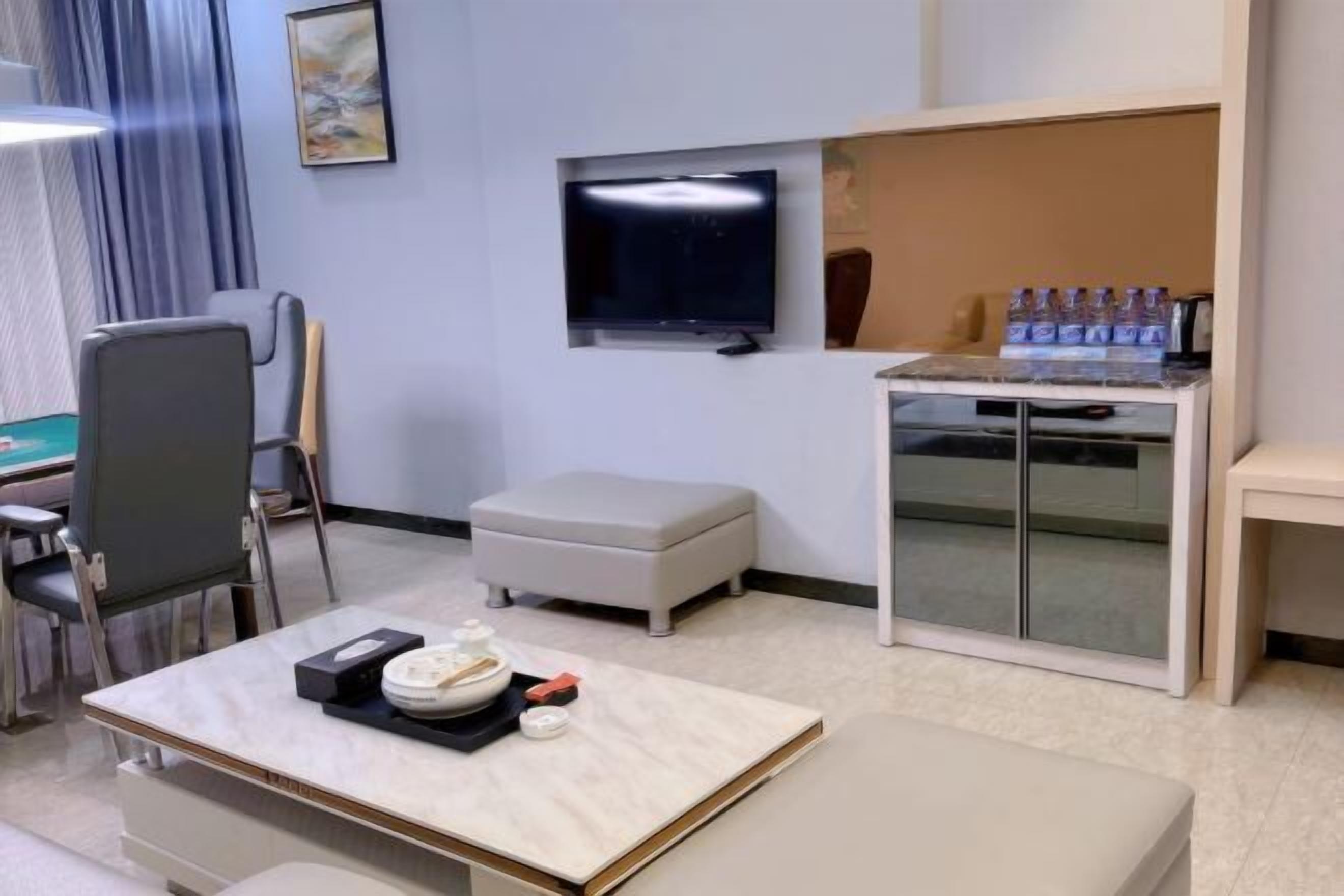 Et hotelværelse med et fladskærms-tv på væggen, et lille bord med skål og bestik, en polstret bænk og et skab med flasker.