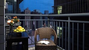 A balcony with a wicker chair, a table with a fruit bowl and flowers, and a cityscape view.
