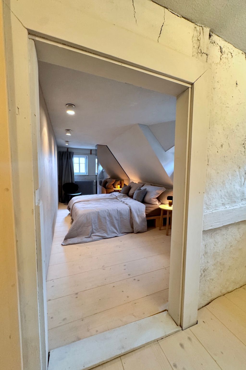 Ein schmaler Flur führt zu einem Schlafzimmer mit schrägverdeckter Decke, einem Bett und einem kleinen Tisch mit einer Lampe.