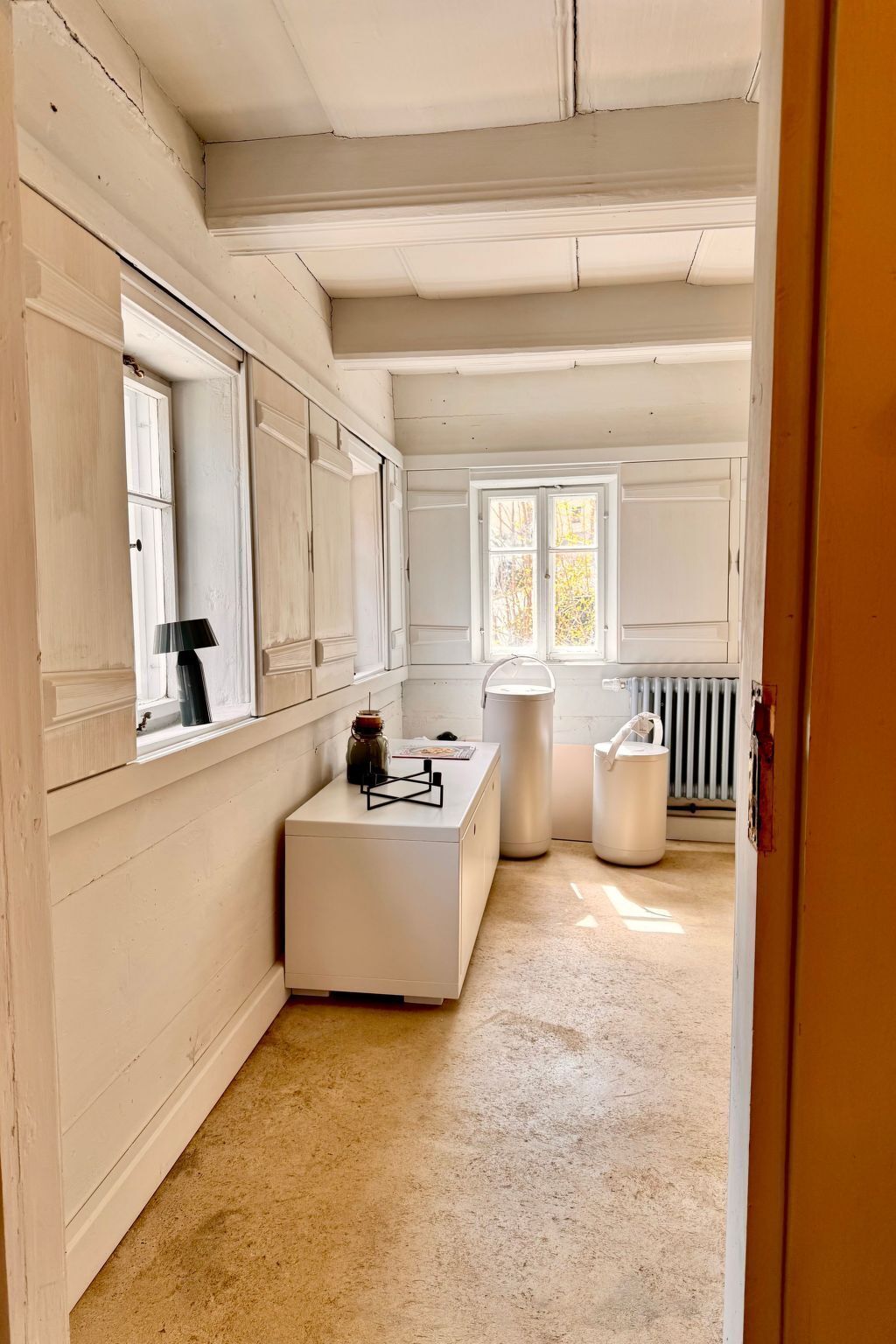A room with a large window, a radiator, and a white cabinet with a lamp.