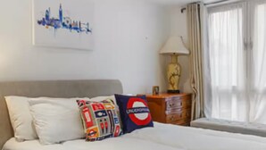 A hotel room with a large bed, a nightstand with a lamp, a cityscape artwork on the wall, and a window with curtains. - Best location! Shoreditch 5 min walk Old St Tube (London)