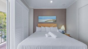 A hotel room with a bed, a nightstand with a lamp, a painting on the wall, and a view of the outdoors through the window. - Spa Haven 18B Top Floor In Room Spa (Airlie Beach)
