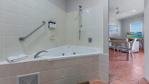 A modern bathroom with a large bathtub, a shower, and a handrail. There is a window with a view of the beach and a dining area with a table and chairs. - Spa Haven 18B Top Floor In Room Spa (Airlie Beach)