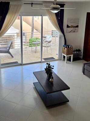 A spacious living area with a sliding glass door to an outdoor patio, a dark wooden coffee table with a plant, and a small white side table with a basket. - Oceanview 2BR + Beachfront Balcony + Sea & Sunset (Punta Cana)