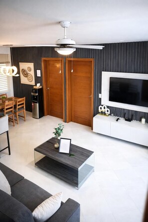A modern living room with a sofa, coffee table, TV, and a ceiling fan. - Oceanview 2BR + Beachfront Balcony + Sea & Sunset (Punta Cana)