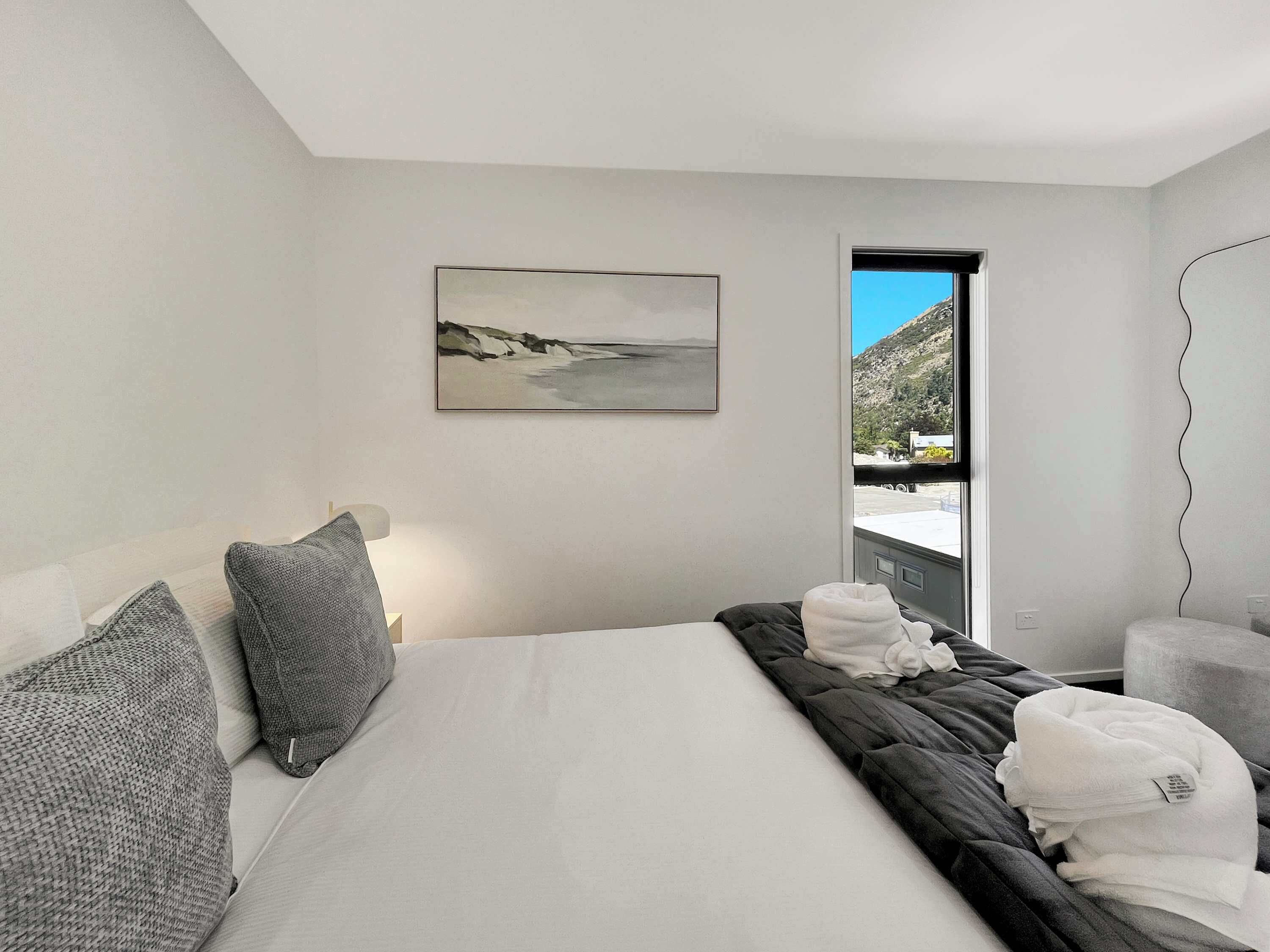 A modern hotel room with a large bed, a painting of a beach, and a view of a mountainous landscape through a window.