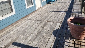 Una terraza de madera con revestimiento azul y ventanas blancas, un patio con bancos azules y una maceta de terracota con follaje.