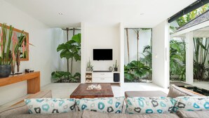 Ein modernes Wohnzimmer mit Flachbildfernseher, Couchtisch aus Holz und einer Couch mit gemusterten Kissen. Auf dem Couchtisch befinden sich große Kübelpflanzen und ein kleines Aquarium.