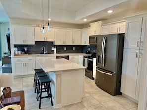 Una cocina moderna con armarios blancos, una isla central con taburetes, electrodomésticos de acero inoxidable y un salpicadero negro.
