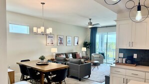 Eine moderne Küche mit weißen Schränken, ein Essbereich mit Holztisch und -stühlen, ein graues Sofa und ein Deckenventilator.