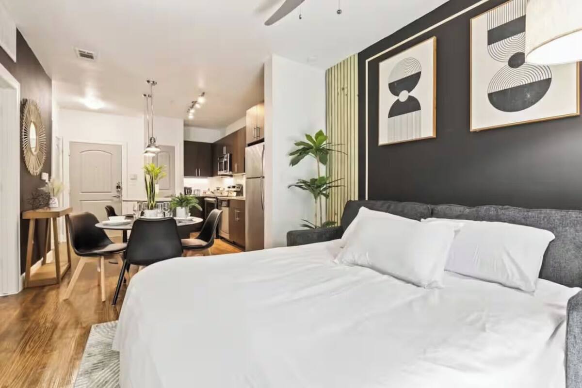 A modern hotel room with a large bed, a dining area, and a kitchen in the background.