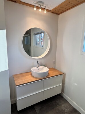 A bathroom with a round mirror, white sink, wooden countertop, and a window with blinds. - Chalet Vous - Mountain view (Beaupré)