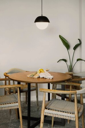 A round wooden table with a black pendant light, surrounded by wicker chairs. - Aposperia Memorable Living K3 (Karpathos)