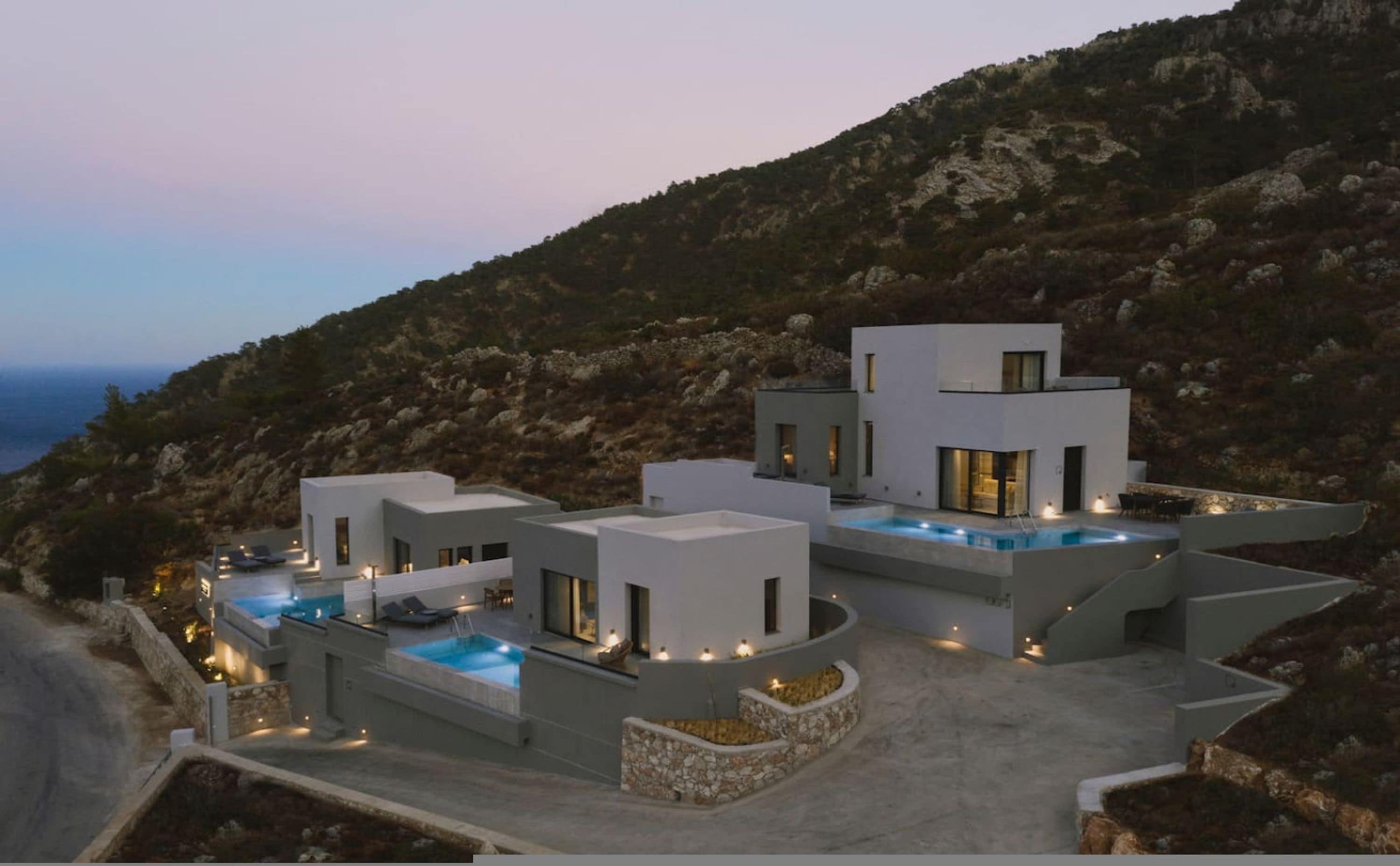A modern multi-story house with a pool, situated on a hillside with a view of the sea.