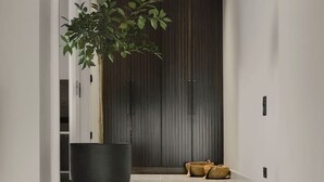 Um interior moderno com uma escada que leva a uma porta de madeira escura, uma grande planta em vaso e um banco com almofada estampada.
