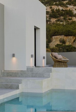 A modern pool area with steps leading to a lounge chair, a white building, and a natural hillside backdrop. - Aposperia Memorable Living K1 (Karpathos)