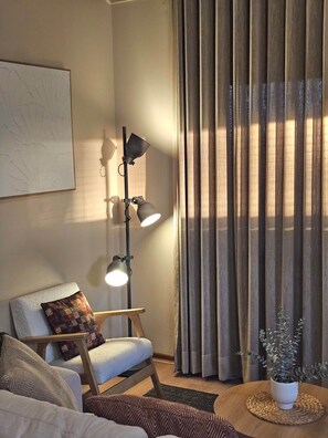Een woonkamer met een bank, een fauteuil, een vloerlamp, een plant en een raam met gordijnen.
