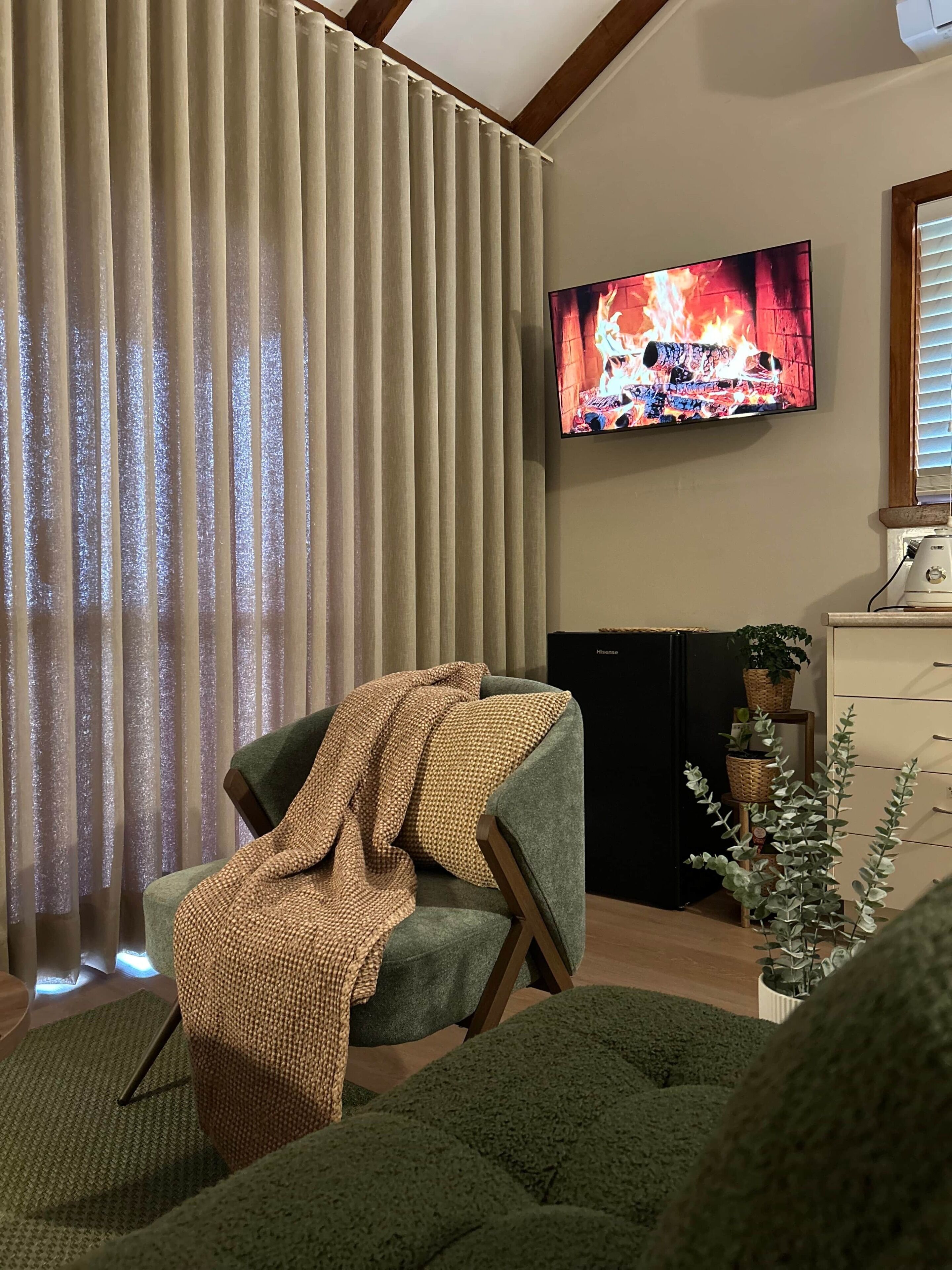 Olohuoneessa on televisio, jossa näkyy kuva takkatulesta, vihreä nojatuoli, sohva ja huonekasvi.