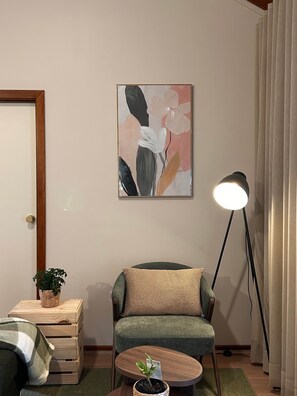 Een woonkamer met een groene fauteuil, een vloerlamp, een schilderij, een kleine plant en een houten bijzettafeltje.