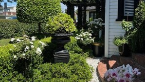 A well-maintained garden with a variety of plants and a brick pathway leading to a white building with black trim. - Escape to La Belle Maison, a Tranquil 3 Bedroom villa in central Tauranga. (Tauranga)
