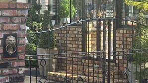 A brick entrance gate with a metal fence, a glass greenhouse structure, and a building with a tiled roof and striped awning. - Escape to La Belle Maison, a Tranquil 3 Bedroom villa in central Tauranga. (Tauranga)