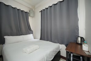 Quarto de hotel com cama, cortinas cinzas, uma mesinha com chaleira e unidade de ar condicionado na parede.