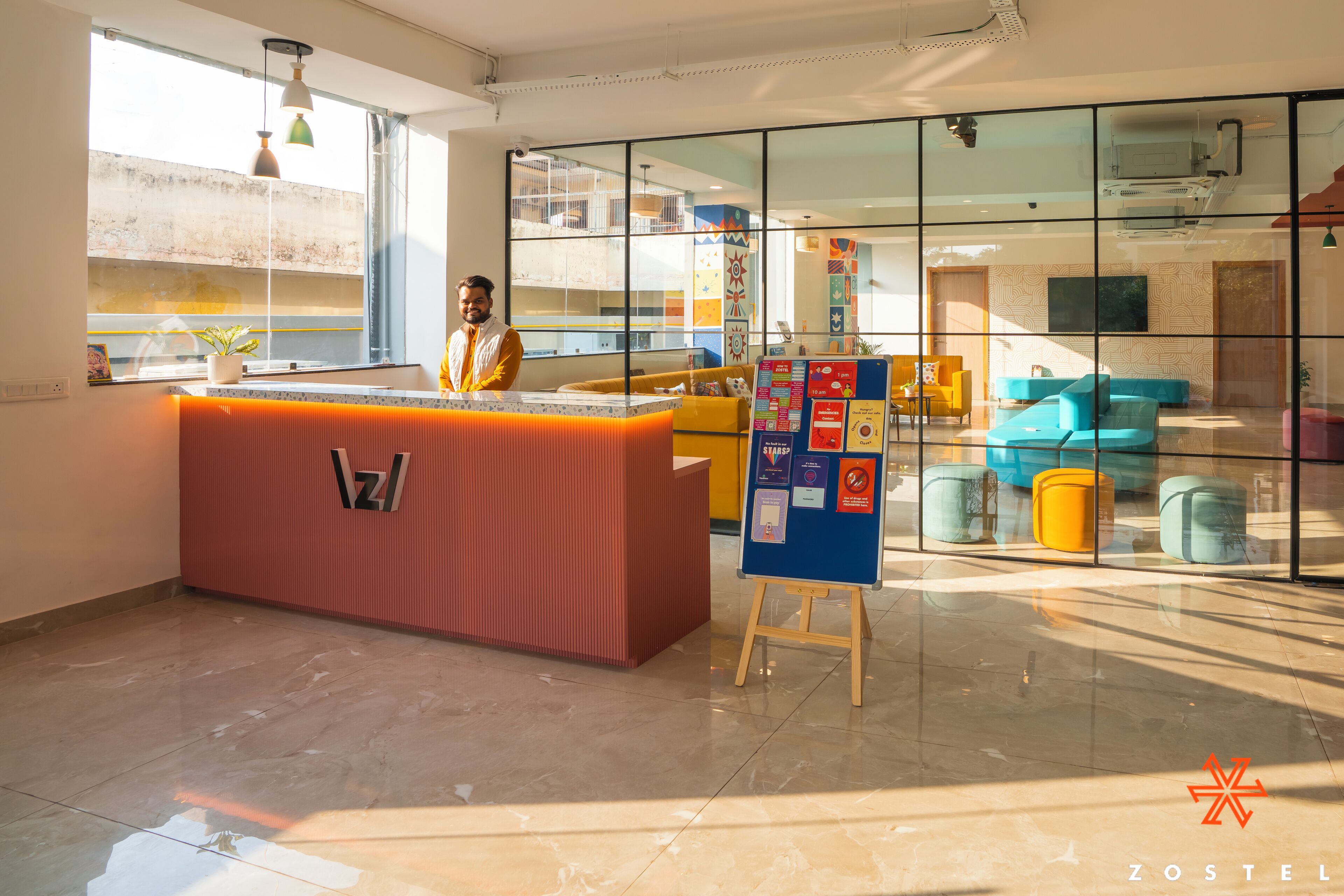 A modern hotel lobby with a reception desk, glass partitions, colorful seating, and a visible staircase.