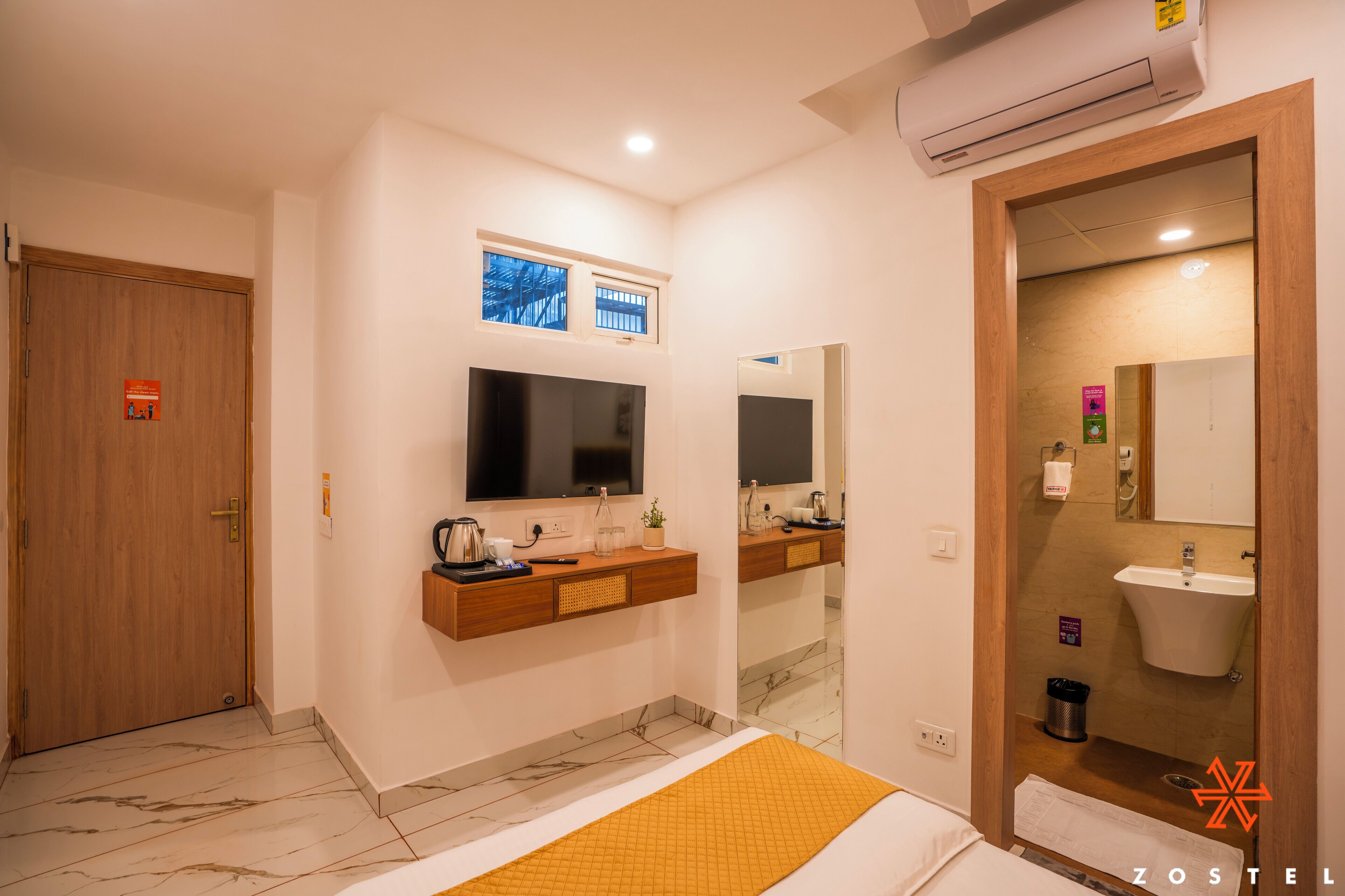 A modern hotel room with a wooden door, a large mirror, a flat-screen TV, a small window, a bathroom with a sink and mirror, and a bed with a yellow blanket.