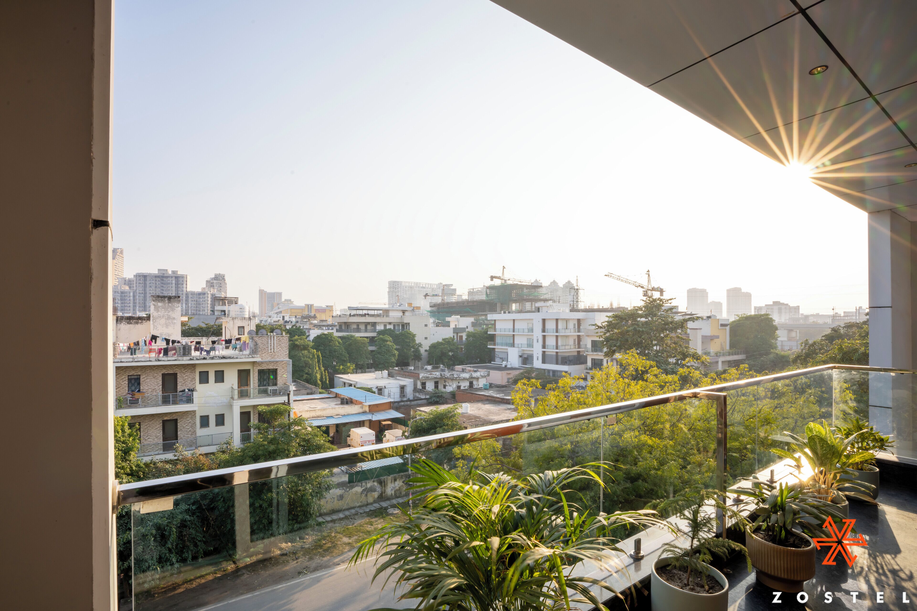 Un balcón con vista a un paisaje urbano, plantas en macetas y el sol brillando a través de la ventana.