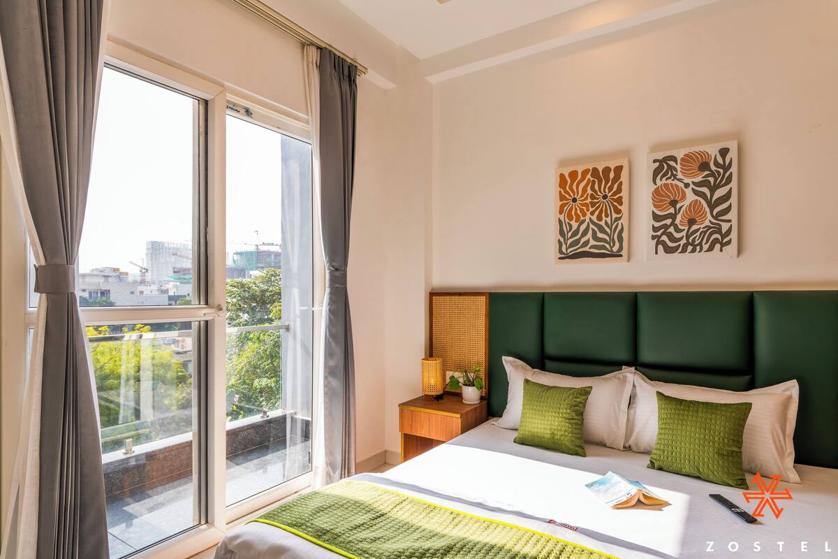Una camera d'albergo con una grande finestra, un letto con biancheria verde e bianca, un comodino con una lampada e una pianta, e due opere d'arte a parete.