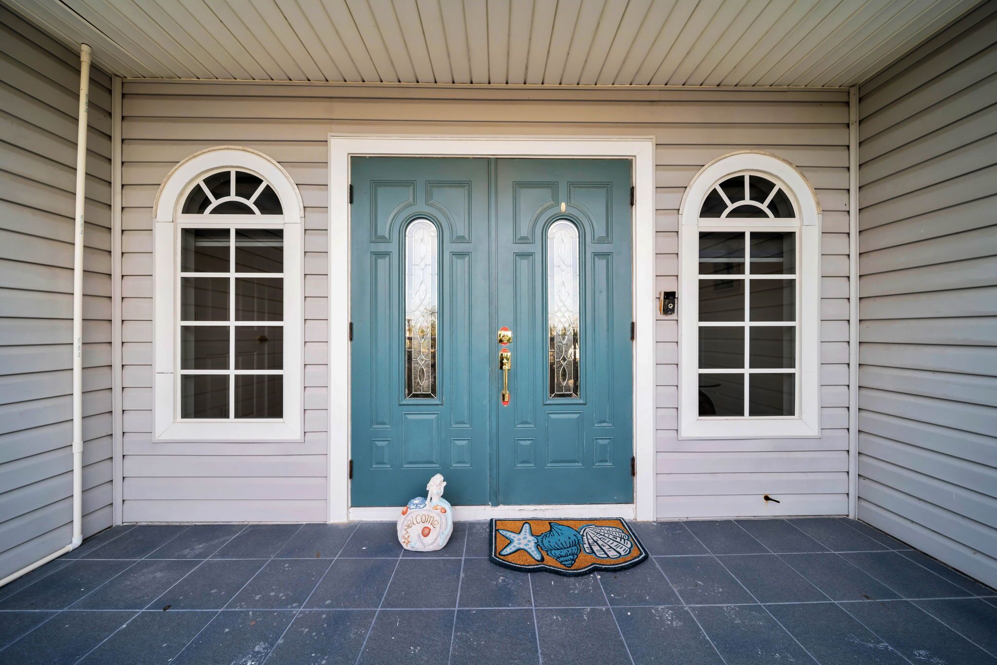 Une maison avec une porte à double battant de couleur turquoise, deux fenêtres en arc de cercle, un paillasson et un tapis d’entrée orné d’un motif d’étoile de mer.