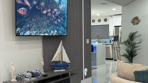 A modern living room with a flat-screen TV mounted on the wall, a built-in fireplace, a curved sofa, and a kitchen area in the background. - Charming Fort Myers Beach Escape 2BR/2BA Near Sand & Sun restaurants and fun. (Fort Myers Beach)