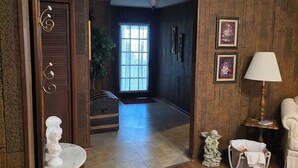 A room with wooden flooring, a wooden door with a decorative handle, a white pedestal table, a lamp, and a small statue.