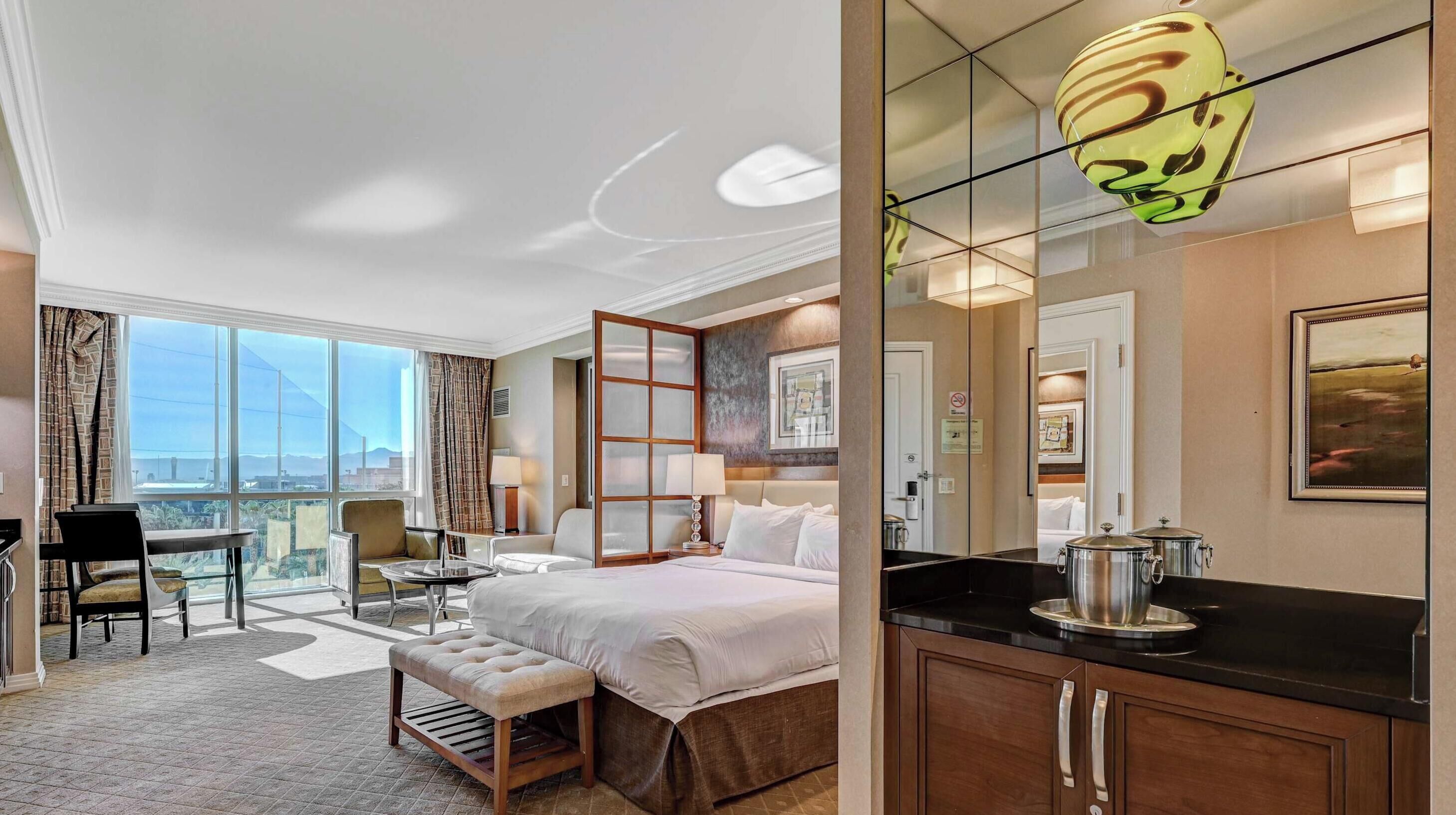 A hotel room with a large bed, a desk, a chair, a small table, a bench, a kitchen area with a stove, and a large window offering a city view.