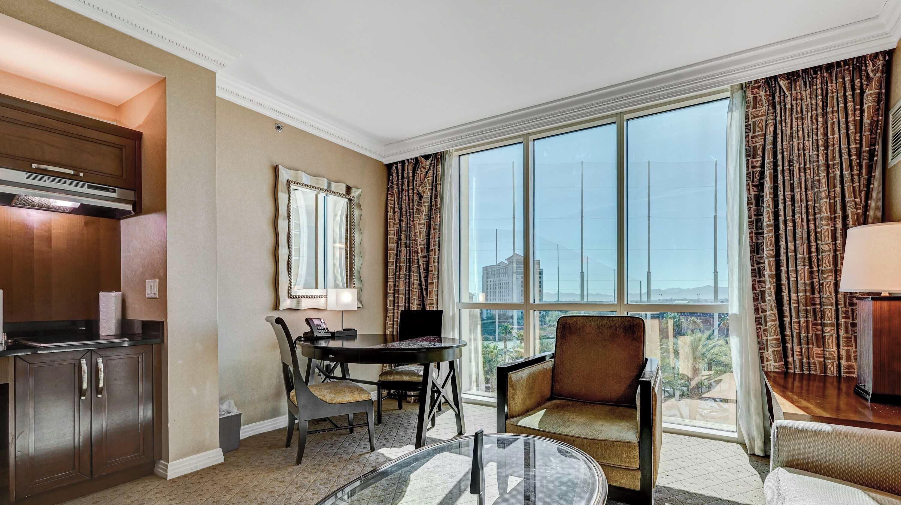 Une chambre d’hôtel équipée d’une table à manger, de chaises, d’un canapé, d’une table en verre, d’un coin cuisine et de grandes fenêtres avec des rideaux.