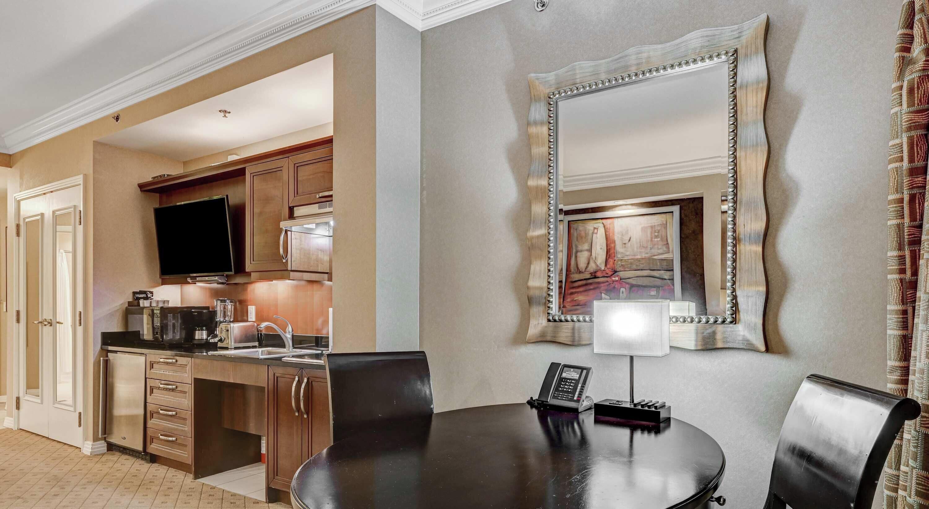 Une chambre d’hôtel équipée d’une table à manger, de chaises, d’un coin cuisine, d’un téléviseur à écran plat et d’un grand miroir.