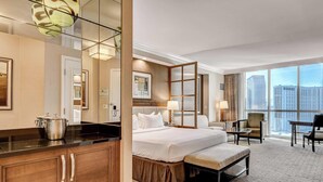 Une chambre d’hôtel comprenant un lit, des tables de chevet, un bureau, des chaises, une grande fenêtre donnant sur la ville et une kitchenette.