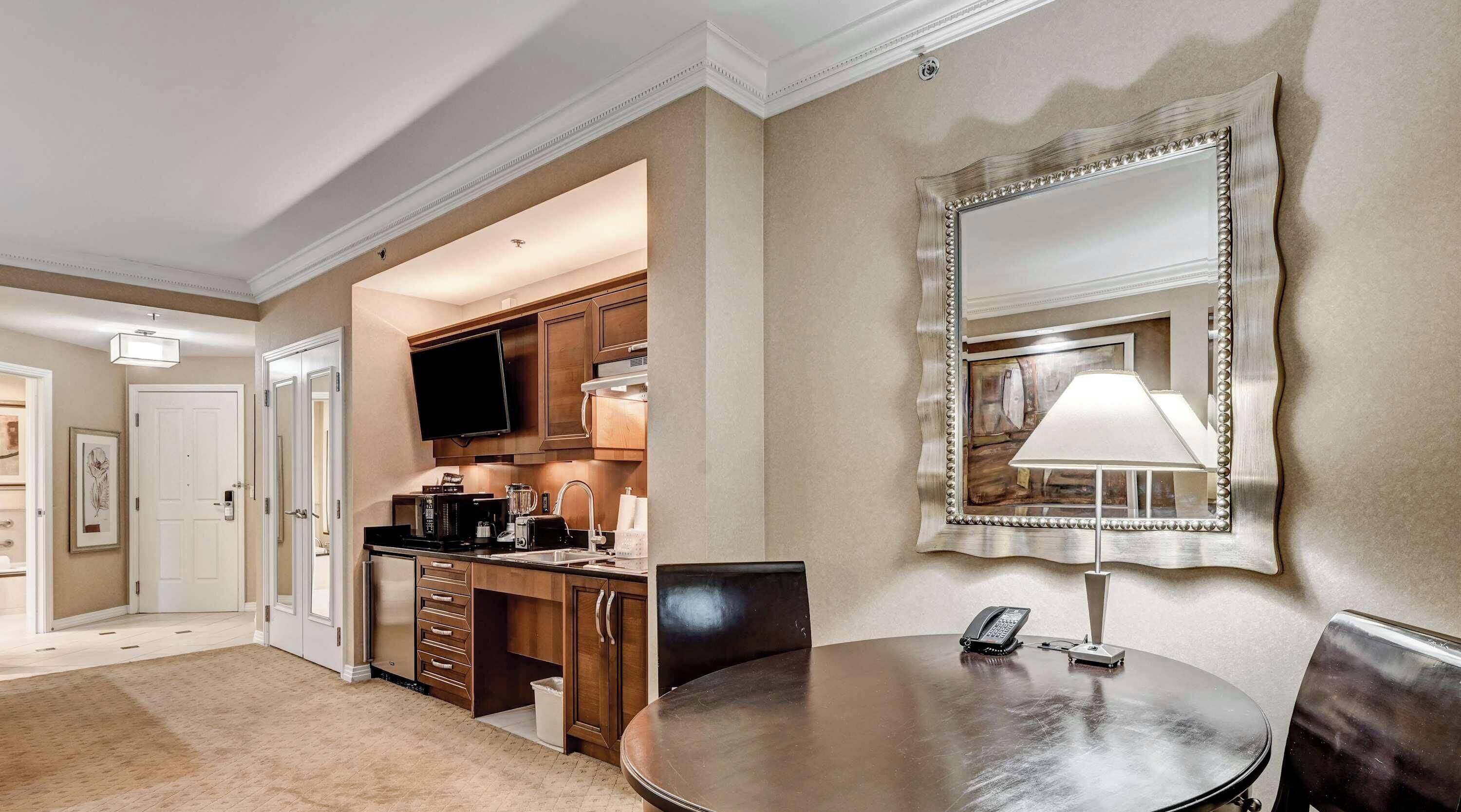 A hotel room with a dining area, a kitchen, a large mirror, and a telephone.